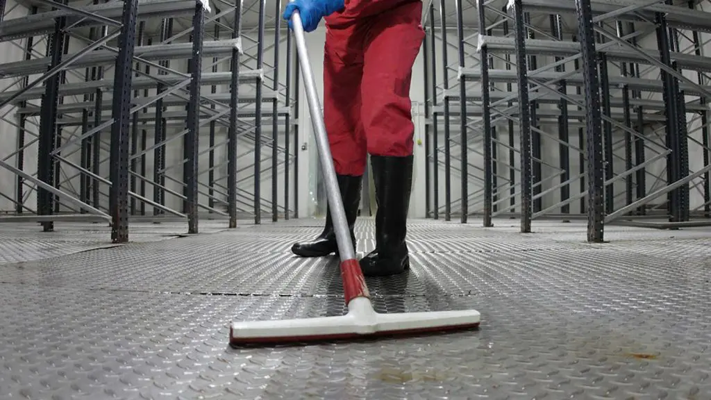 Persona en traje rojo y negro, sobre un suelo metálico, con un trapeador en la mano realizando limpieza industrial.
