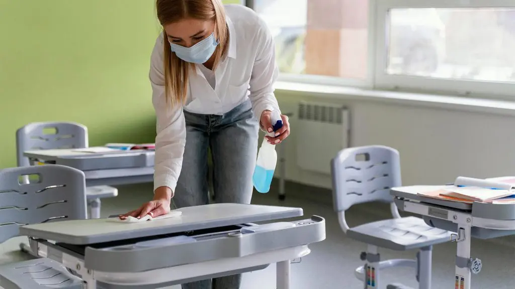 Una mujer con mascarilla desinfectando escritorios en un aula, realizando el servicio de limpieza escolar.