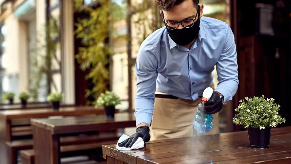 Hombre con mascarilla y guantes limpiando una mesa de un restaurante, asegurando un ambiente higiénico y seguro.