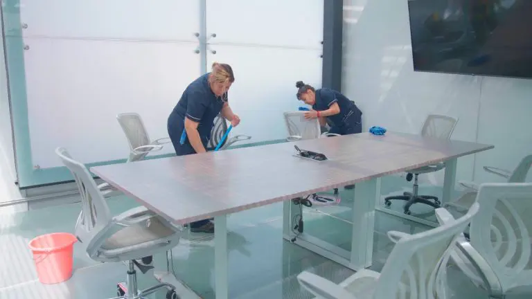 Dos mujeres organizando y limpiando una mesa de conferencias en una oficina, contribuyendo a un espacio de trabajo eficiente.