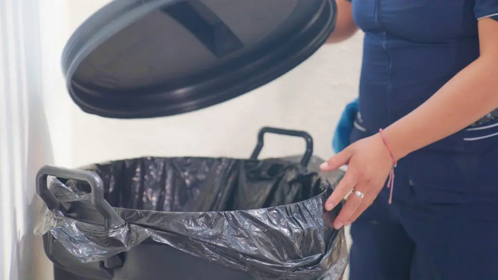Una mujer con uniforme azul coloca un bote de basura negro en una habitación.