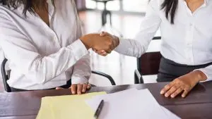 Bolsa de trabajo Dos mujeres de negocios estrechándose la mano en una mesa, simbolizando un acuerdo laboral.
